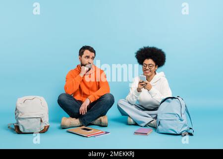 cheerful african american student using smartphone near offended boyfriend sitting on blue background Stock Photo