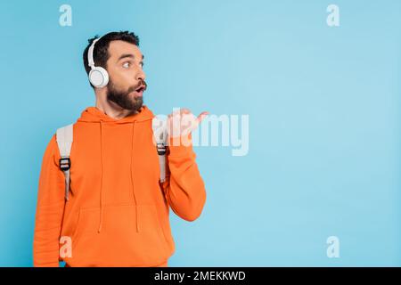 astonished student in wireless headphones and orange hoodie looking away and pointing with thumb isolated on blue Stock Photo