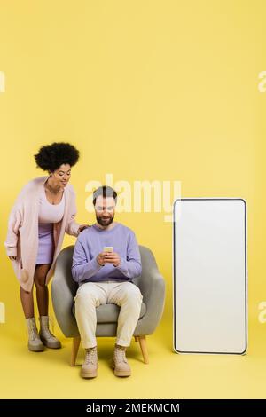 full length of man using smartphone in armchair near smiling african american woman and huge phone template on yellow background Stock Photo