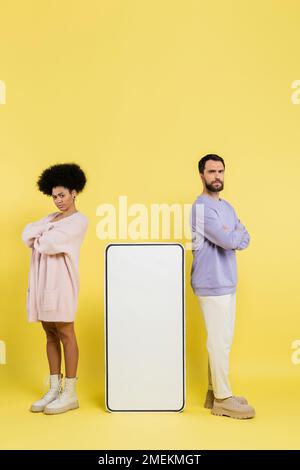 full length of displeased interracial couple standing with crossed arms near huge phone template on yellow background Stock Photo