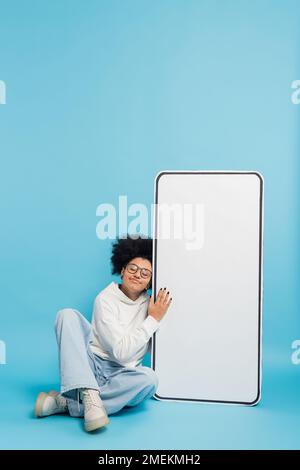 curly african american woman in eyeglasses and hoodie leaning on phone template while sitting with closed eyes on blue background Stock Photo