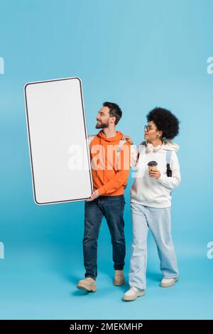 bearded student carrying mobile phone template near african american girlfriend with coffee to go on blue background Stock Photo