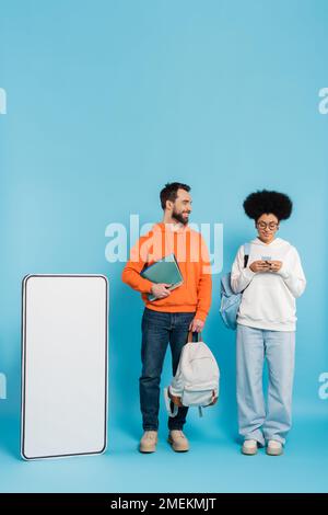 african american woman using smartphone near bearded student and white phone template on blue background Stock Photo