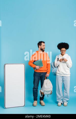 african american woman using smartpone near bearded student and white phone template on blue background Stock Photo
