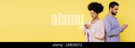 stylish interracial couple standing back to back and chatting on cellphones isolated on yellow, banner Stock Photo