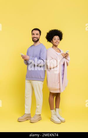 full length of happy and stylish multiethnic couple looking at camera while standing with cellphones on yellow Stock Photo