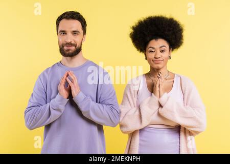cunning interracial couple smiling at camera and showing please gesture isolated on yellow Stock Photo
