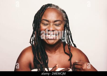 Cheerful mature woman laughing happily while wrapped in a bath towel. Refreshed woman with dreadlocks standing against a white background. Joyful midd Stock Photo