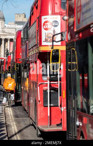 Just Eat delivery rider squeezing down the side of a queue of red London buses in a bus lane in Whitehall, Westminster, London, UK. Cyclist on inside Stock Photo