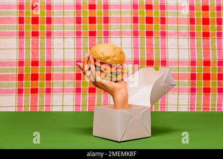 Food pop art photography. Female hand sticking out box and holding burger on green tablecloth. Junk food aesthetics Stock Photo
