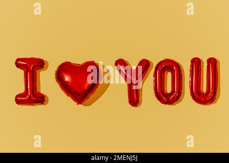 a red heart-shaped balloon and some letter-shaped balloons forming the sentence I love you on a yellow background Stock Photo