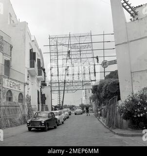 1960s, historcial, workers constructing a giant outdoor display as part of a festival celebration across a street in Ischia town, Italy. Italian cars of the era parked in the street, including several small Fiat's and a Lancia saloon, with a numberplate, 631257 Roma. Beside the cars, on the left a building with a sign 'Terme' (Spa), perhaps the Hotel La Villarosa Terme, a villa turned into a hotel in the 1950s. Stock Photo