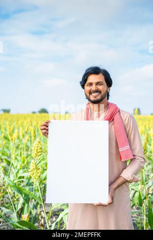 Vertical shot of happy young indian farmer holding empty sign board at agricultural farmland - concept of advertising, promotion and modern farming Stock Photo