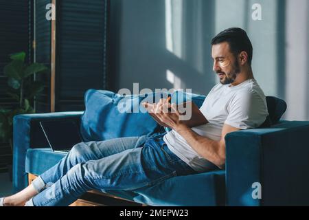 Successful bearded young man writing notes in notebook while sitting at home in living room on the sofa in casual clothes. Writing daily routine in Stock Photo