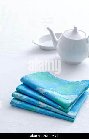 Pile of folded marbled napkins, teapot and bowl in background, close-up Stock Photo