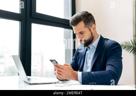 Focused serious caucasian business man, manager, director, sits at a desk in the office, using his smartphone, chatting with a colleague, reading information, answering an email, making an appointment Stock Photo