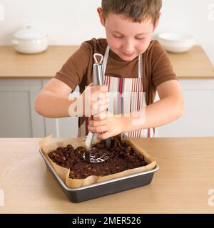Boy using masher to press chocolate and biscuit fridge cake into cake tin, 8 years Stock Photo