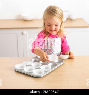Girl putting cake cases onto cake tin, 3 years Stock Photo