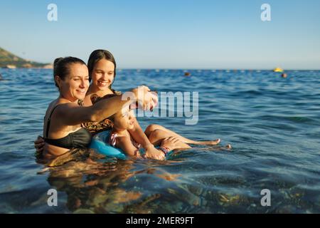 A happy young mother and her two cheerful daughters in bright swimsuits with a blue small inflatable circle take photos for memory, in the azure summe Stock Photo