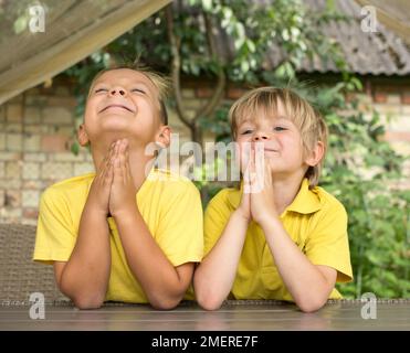 two happy boys of 5 years old in yellow t-shirts pray to God with their hands together, their faces raised to the sky. Faith in the best, hope, dreams Stock Photo