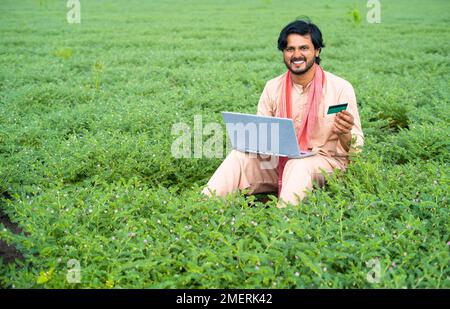 wide shot of Young farmer at field making online payment using credit card on laptop - concept of modern farming, online banking service and wireless Stock Photo