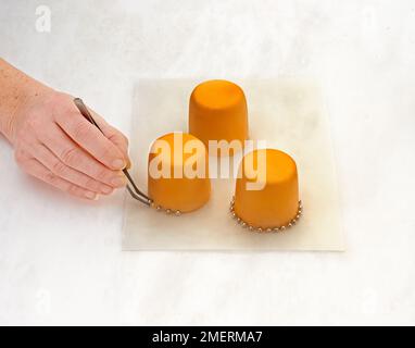 Sticking small silver edible balls at the bottom of three small cakes covered with yellow icing Stock Photo
