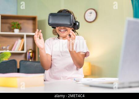 happy smiling teenager Girl kid with vr or virtual head set experiencing online or virtual class using hand gesture in front of study table at home - Stock Photo