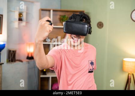 sweet excited young man with VR or virtual reality goggles doing workout by using joystick at home - concept of metaverse, health care and technology. Stock Photo