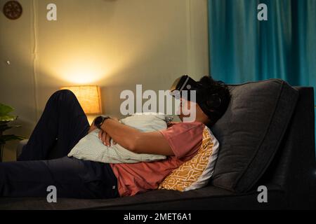 Tired young man with VR or virtual reality goggles sleeping on couch at home during night - conept of addiction, relaxation and futuristic Stock Photo