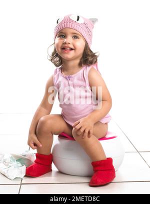 Girl wearing pink vest and pink wool hat, red slippers sitting on white potty, nappy on floor, 20 months Stock Photo