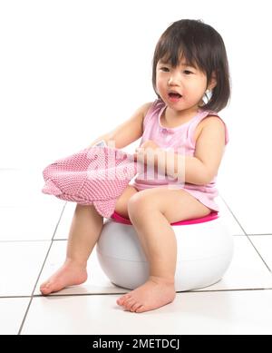 Girl wearing pink vest sitting on white potty, holding pink woolly hat, 20 months Stock Photo