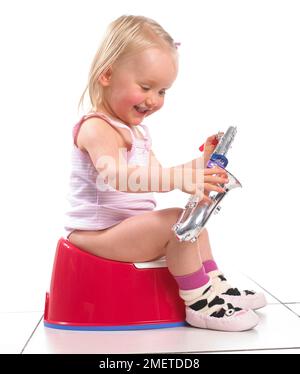 Girl wearing vest and slippers sitting on a red potty playing with a toy saxophone, 20 months Stock Photo