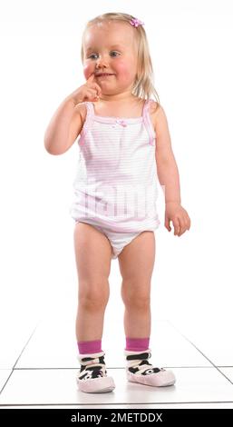 Girl wearing vest, pants and slippers, holding finger to her mouth, 20 months Stock Photo