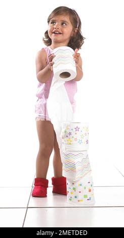 Girl wearing vest, pants and slippers making a tower from rolls of toilet paper, 20 months Stock Photo