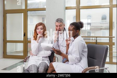 A group of doctors in a medical clinic sit on chairs and discuss the patient's diagnosis with surprise Stock Photo