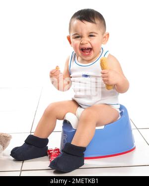Boy wearing vest and slippers sitting on blue potty holding bread, 15 months Stock Photo