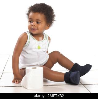 Boy wearing vest and slippers sitting on floor with roll of toilet paper, 17 months Stock Photo