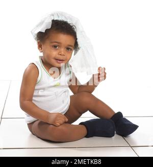 Boy wearing vest and slippers sitting on floor with nappy on head, 17 months Stock Photo