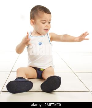 Boy wearing vest, pants and slippers, sitting on the floor, 15 months Stock Photo
