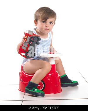 Boy sitting on a potty shaped as a car with steering wheel holding toy fire engine, 20 months Stock Photo