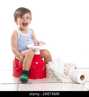Boy sitting on a potty shaped as a car with steering wheel holding toilet roll, 20 months Stock Photo