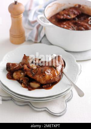 Coq au vin, chicken legs with shallots, mushrooms, rosemary and thyme in red wine sauce, served on plate, pan and pepper mill in background Stock Photo