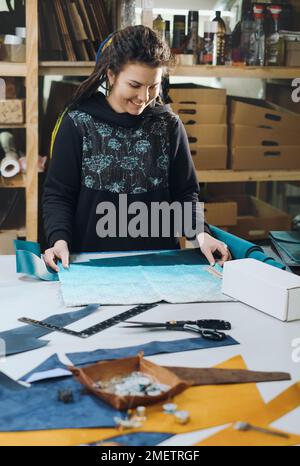 Seamstress at work. Dressmaker making clothes and bags in workshop. Tailor marking fabric. Woman working at table with cut textile, thread, pins Stock Photo