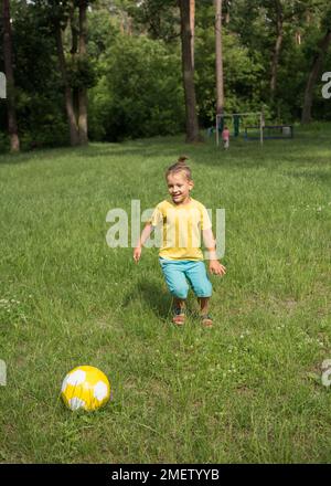 boy of 5-6 years old runs in the park on green grass and kicks a yellow soccer ball. Active lifestyle, fun activities for children in the summer outdo Stock Photo