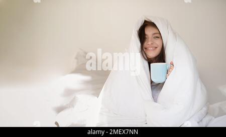 Cheerful young european woman wrapped in white blanket sits on white bed with cup of coffee Stock Photo
