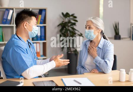Serious mature korean doctor in protective mask consults old european female patient in clinic Stock Photo