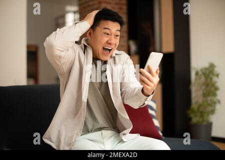Big win. Overjoyed asian man holding smartphone and touching head in joy and excitement, sitting on sofa at home Stock Photo