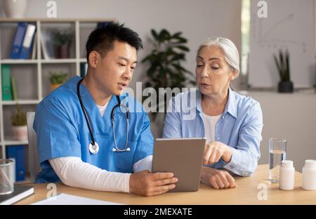 Serious middle aged korean male doctor showing tablet and consulting senior european lady patient in clinic Stock Photo