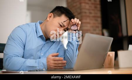 Boring tired mature asian businessman working on laptop computer, sitting at table at home office, panorama Stock Photo