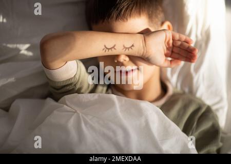 Cheerful cute tired little boy lies on bed puts his hand to head on hand drawn closed eyes in bedroom interior Stock Photo